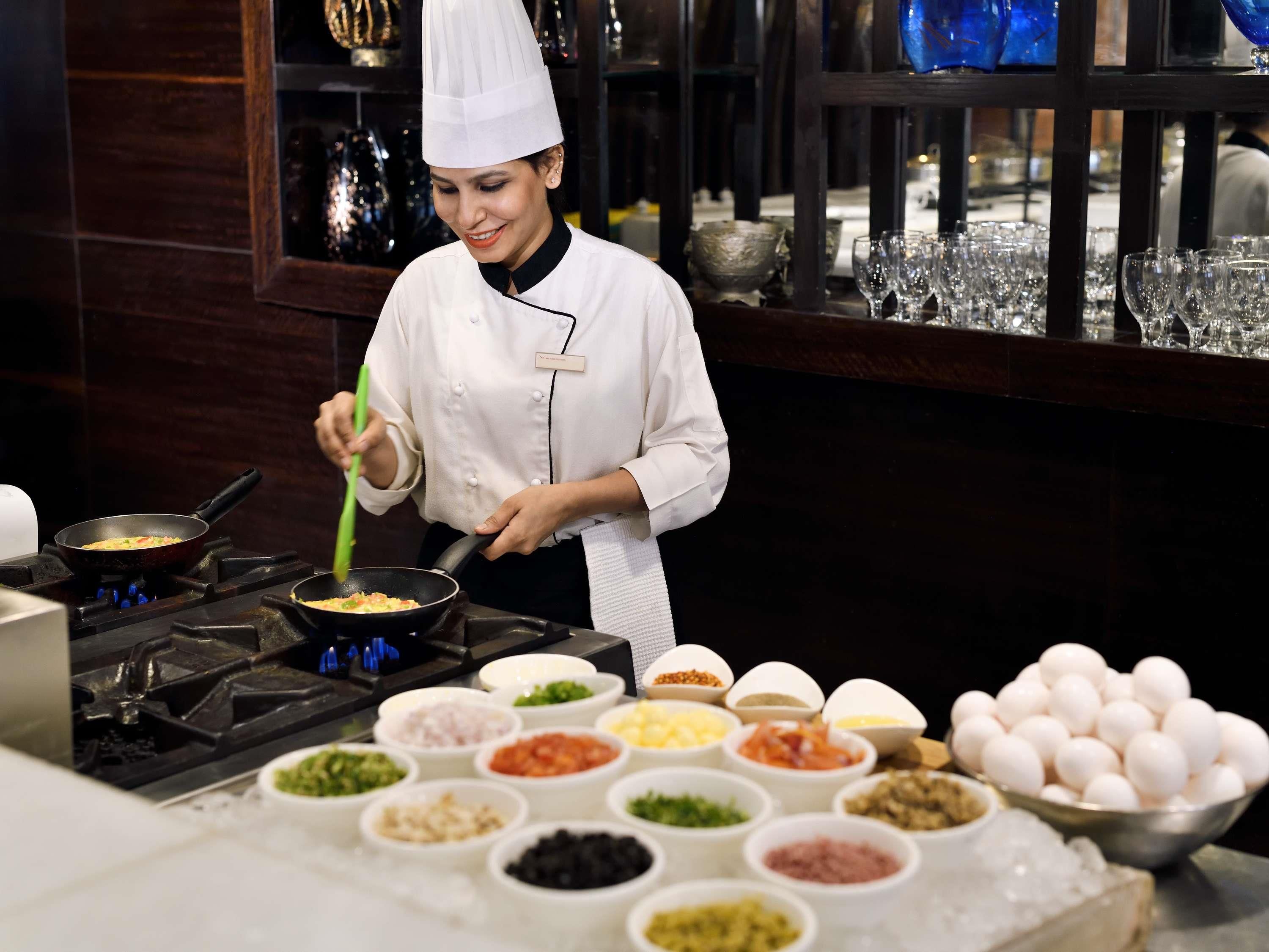 Un chef vêtu d’une blouse blanche prépare un plat dans une cuisine où l’on peut voir divers ingrédients et ustensiles.