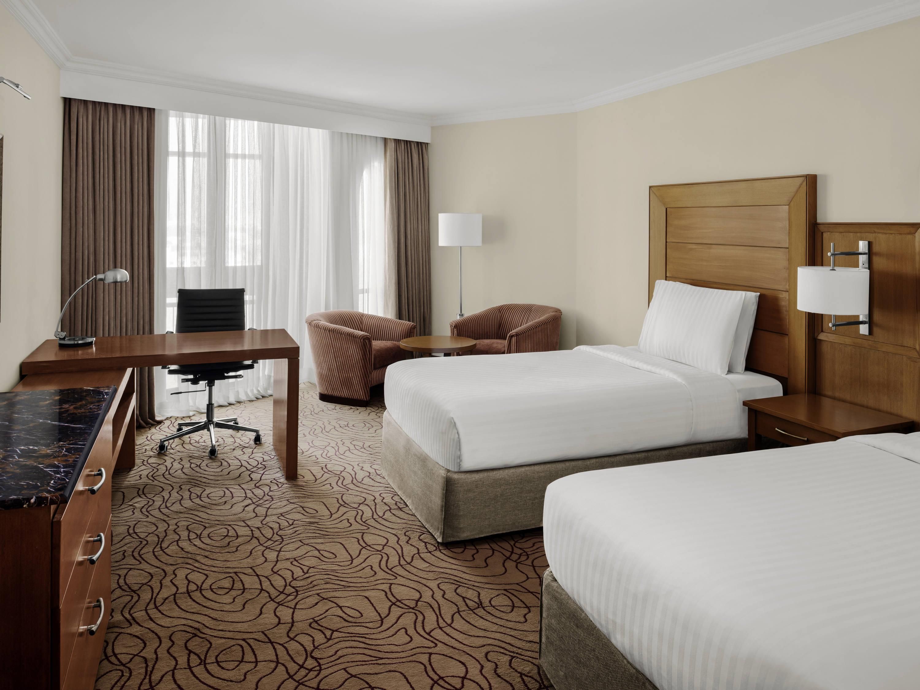 A hotel room with two beds, a desk, a chair, a lamp, and a nightstand.