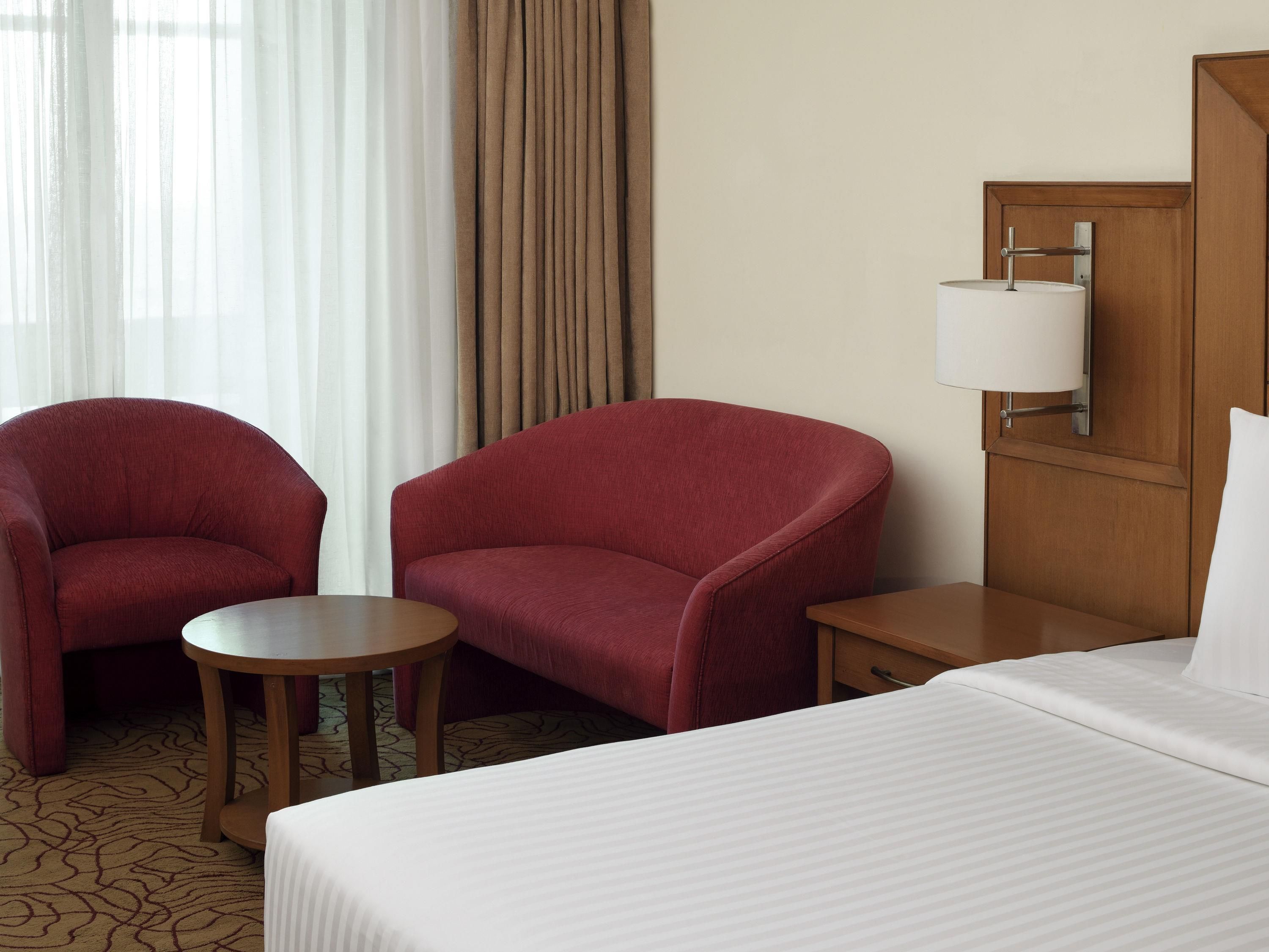 Habitación de hotel con cama, dos sillones, una mesita, una mesita de noche, una lámpara y una ventana con cortinas.