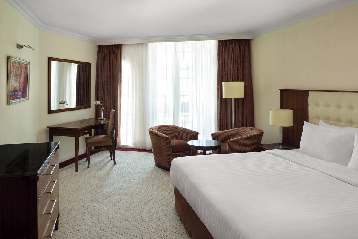 Ein Hotelzimmer mit einem großen Bett, einem Schreibtisch mit Stuhl, einem kleinen runden Tisch und zwei Sesseln.