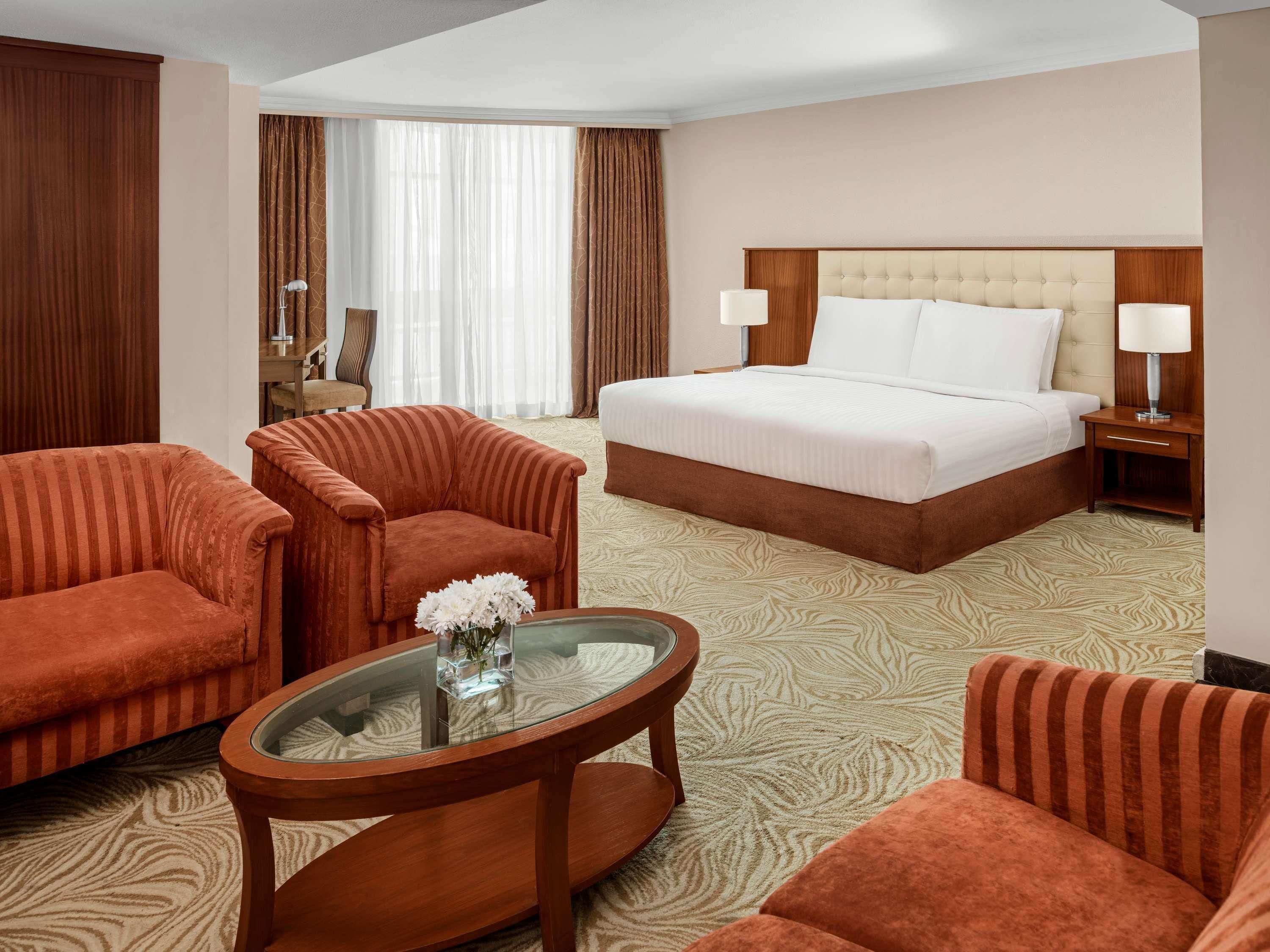 A hotel room with a large bed, two armchairs, a glass coffee table, and a view of the outside.