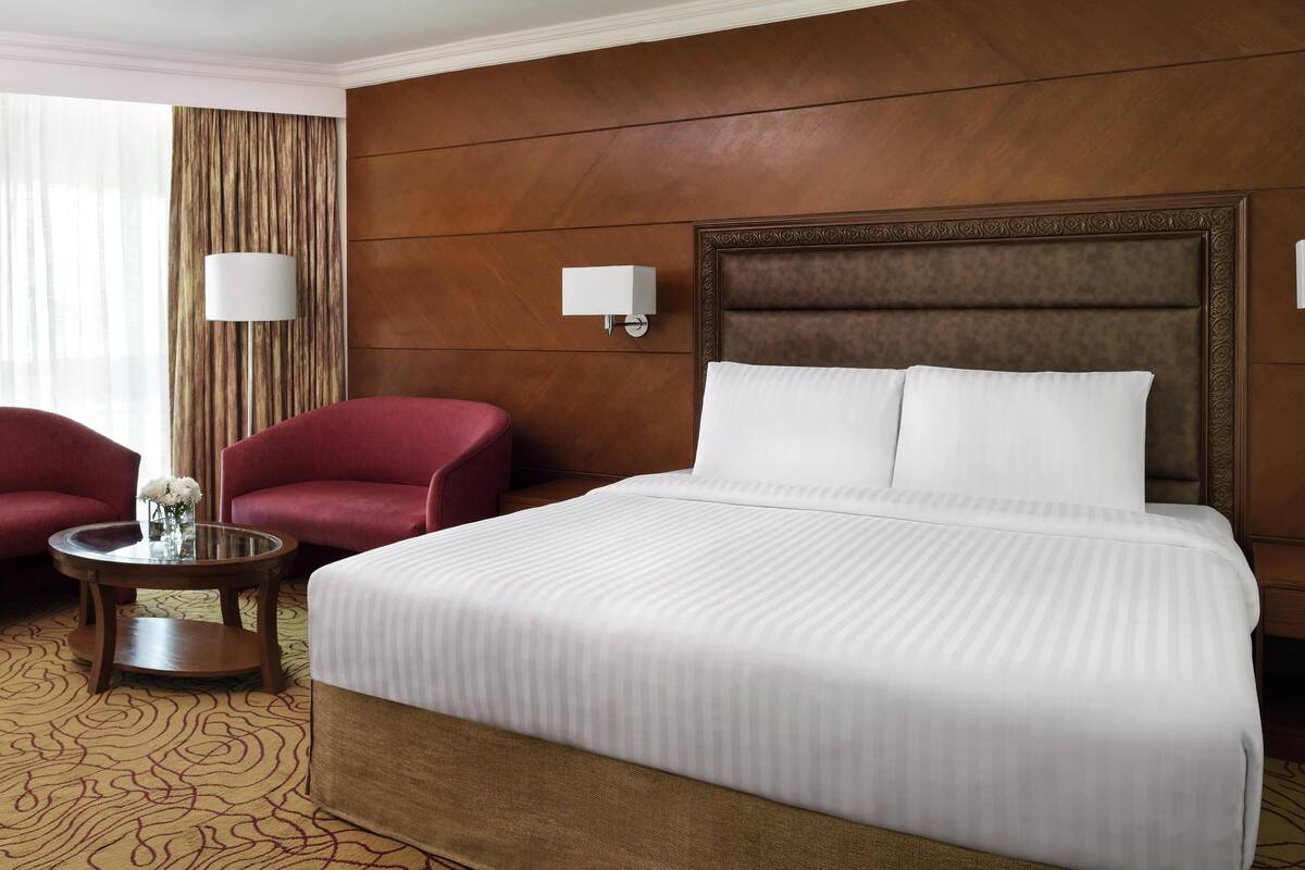 Ein Hotelzimmer mit einem großen Bett, zwei roten Sessel, einem kleinen Tisch mit Blumenvase und einem gemusterten Teppich.