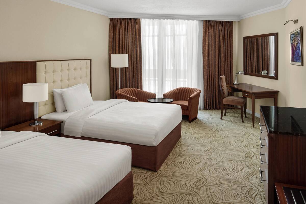 Ein Hotelzimmer mit zwei Betten, einem Schreibtisch, einem Sessel, einem Fernseher und einem großen Fenster mit Vorhängen.