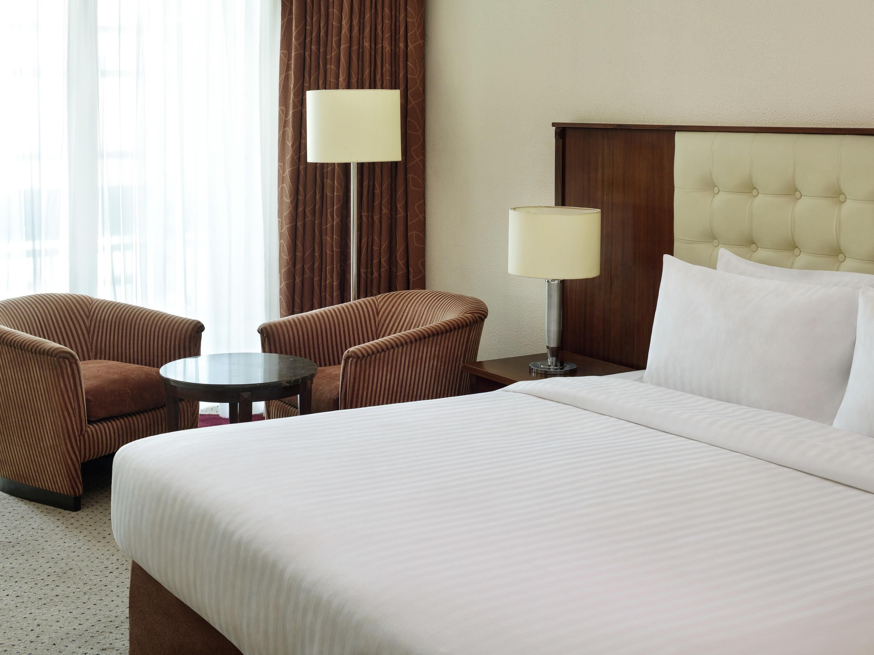 A hotel room with a large bed, two armchairs, a small round table, and two bedside lamps.