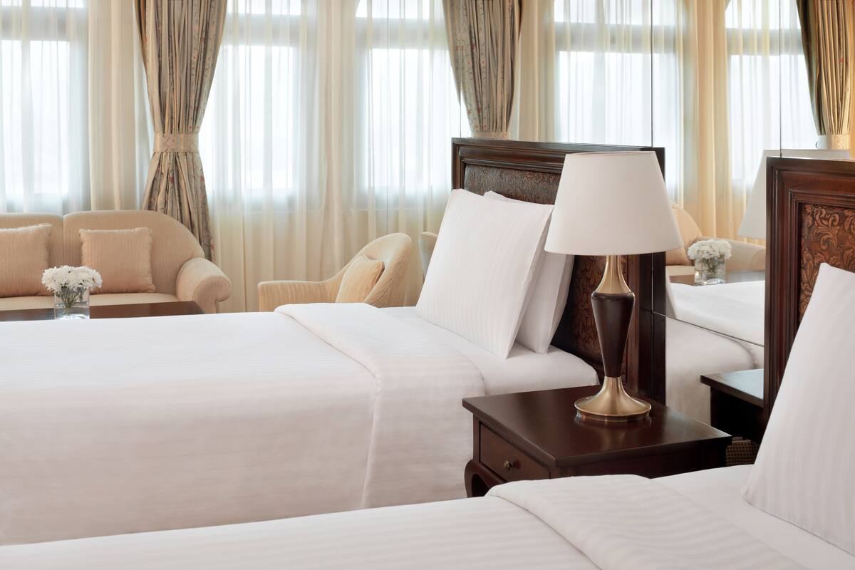 Ein Hotelzimmer mit zwei Betten, einer Sitzecke inklusive Sofa und Sesseln, einem Nachttisch mit Lampe sowie einem Fenster mit Vorhängen.
