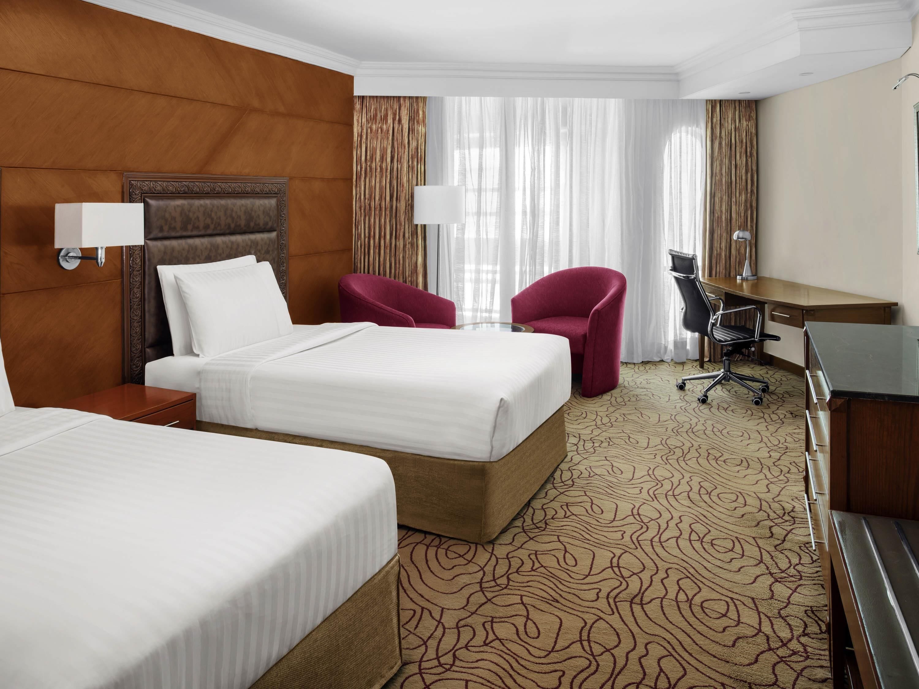 A hotel room with two beds, a desk, a chair, and a large window with curtains.