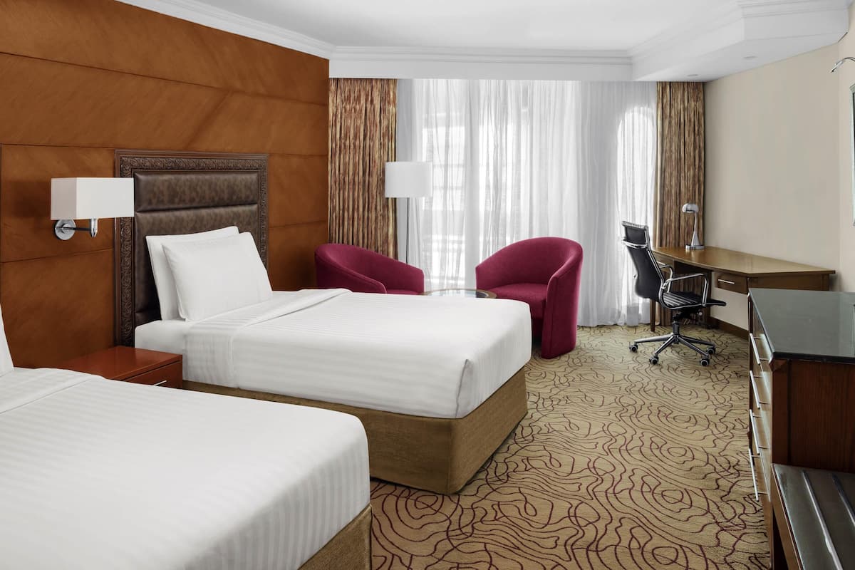 Ein Hotelzimmer mit zwei Betten, einem Schreibtisch, einem Stuhl und einem großen Fenster mit Vorhängen.