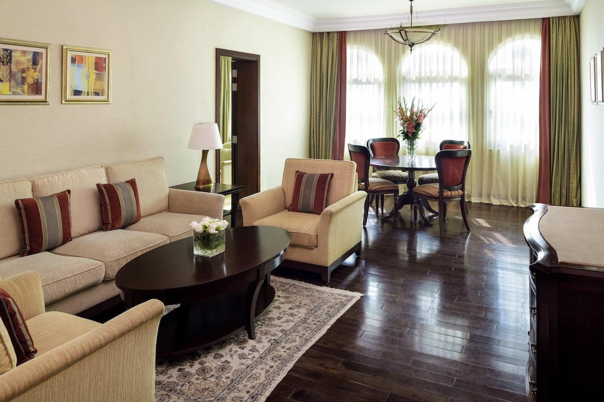A living room with a sofa, armchairs, a coffee table, and a dining table with chairs. There is a lamp, a vase with flowers, and curtains on the windows.