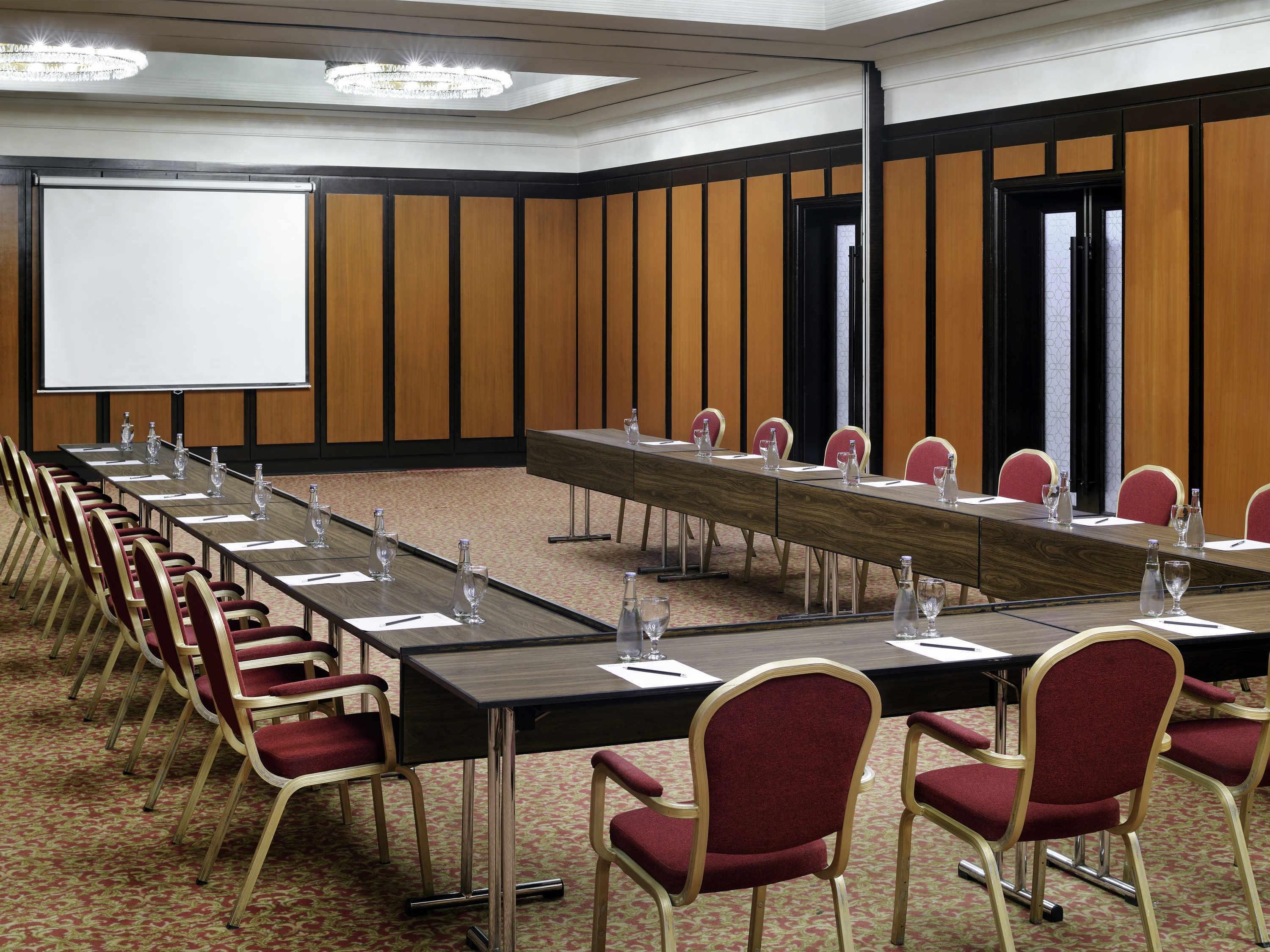Une salle de conférence avec une longue table, des chaises rouges, un écran de projection et un lustre.