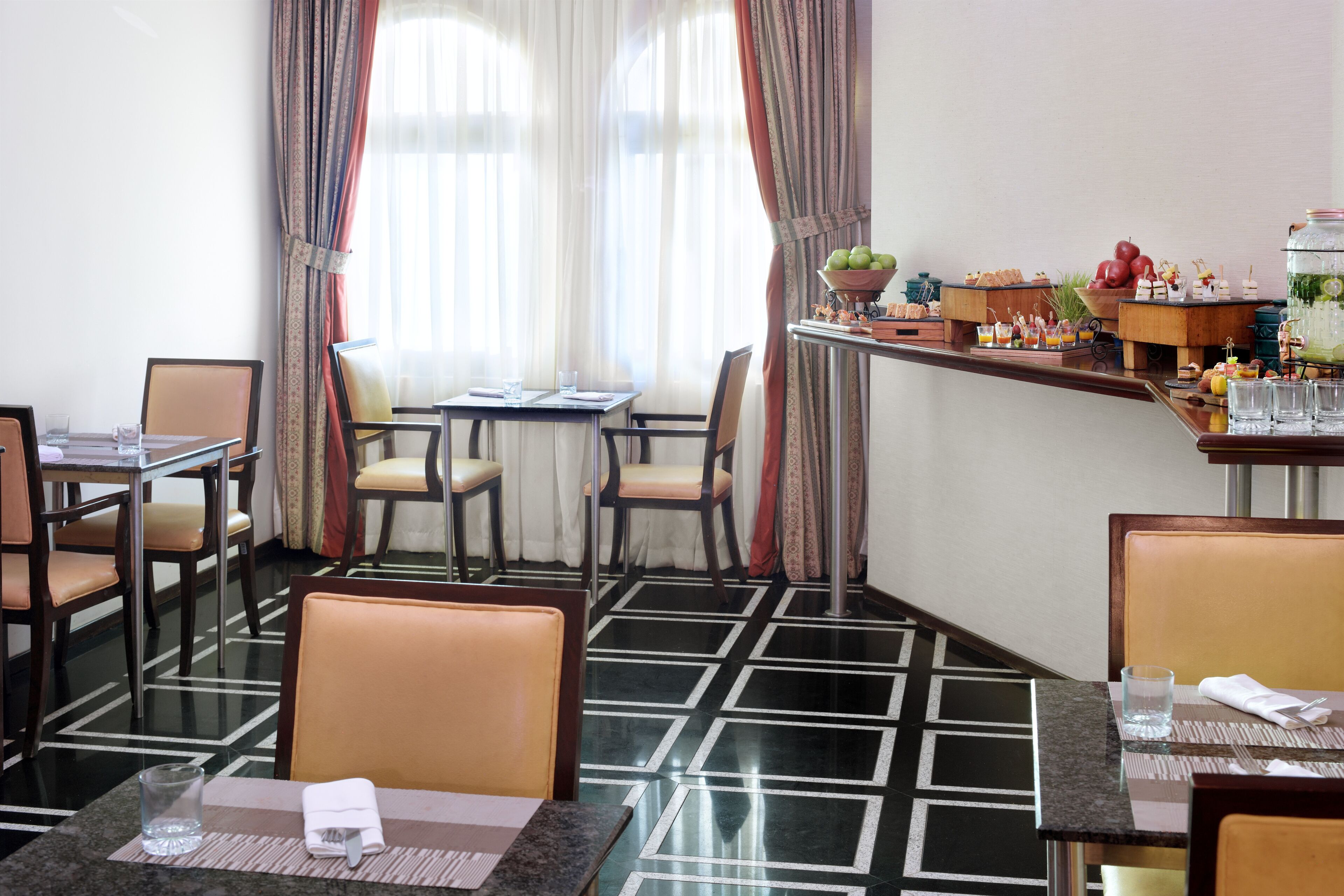 Une salle à manger d’hôtel avec une table dressée pour deux personnes, un buffet proposant divers plats et un bar équipé d’un distributeur de boissons.