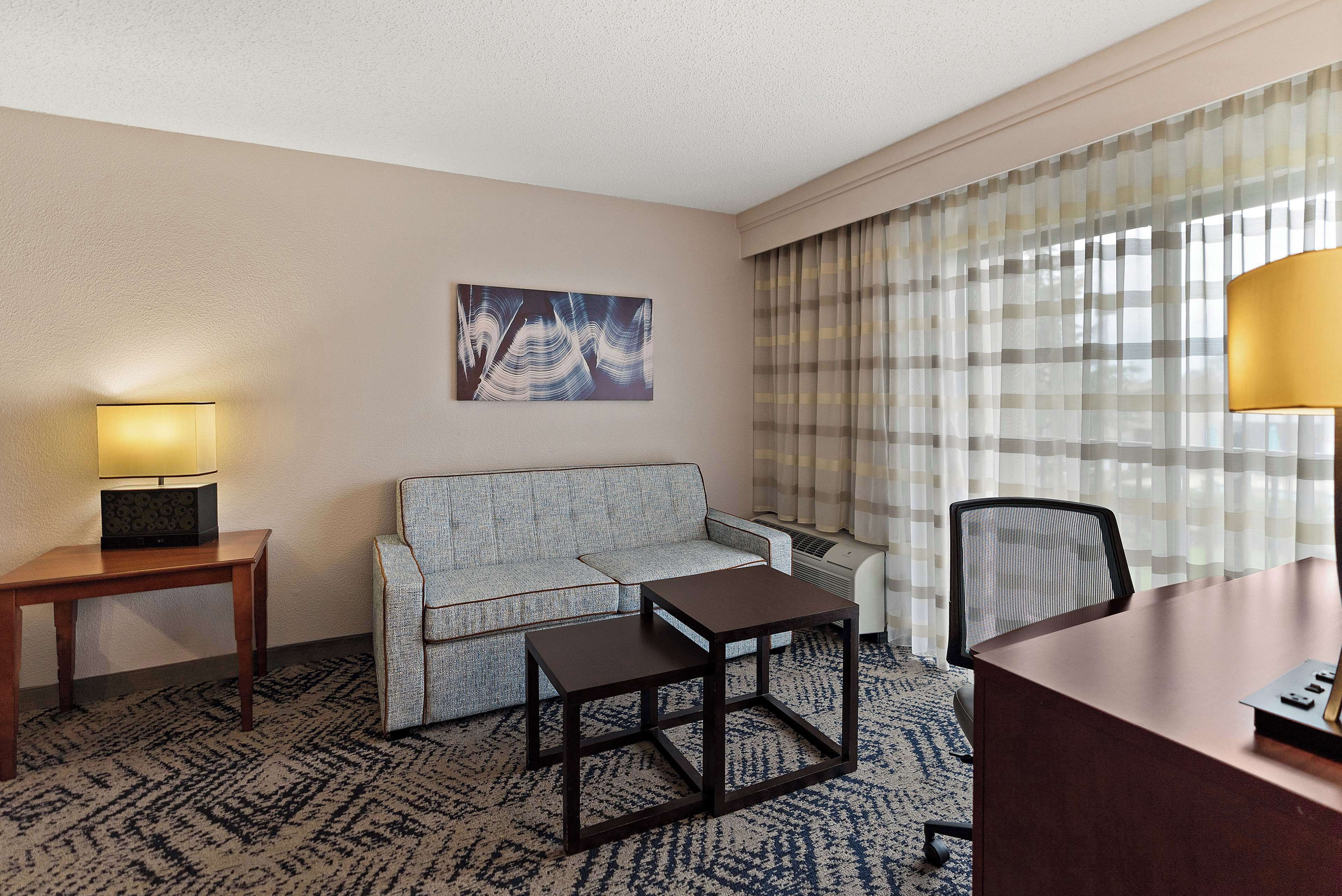 A hotel room with a sofa, a small table, a desk, a lamp, a painting, and a window with curtains.