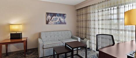 A hotel room with a sofa, a small table, a desk, a lamp, a painting, and a window with curtains.