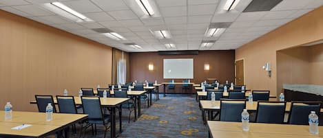 A conference room with multiple tables and chairs, a projector screen, and water bottles on each table.