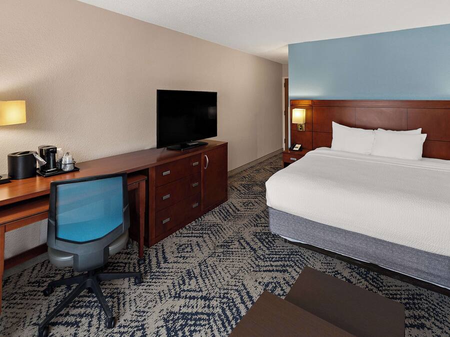 A hotel room with a bed, a desk with a television, a chair, and a nightstand with a lamp.
