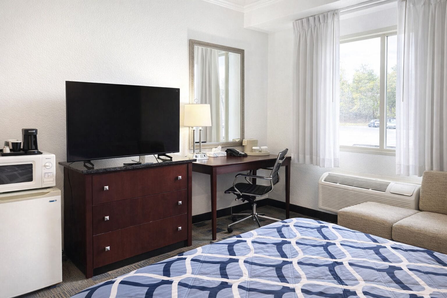 A hotel room with a bed, a desk with a chair, a television, a microwave, a dresser, and a window with curtains.