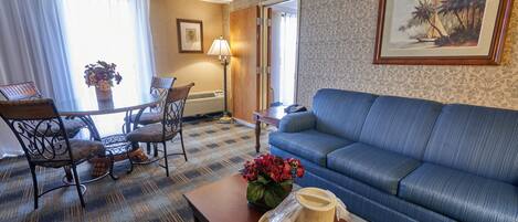 Una habitación de hotel con un sofá azul, una mesa de comedor con sillas, una mesa de centro con un jarrón de flores y un cuadro en la pared.