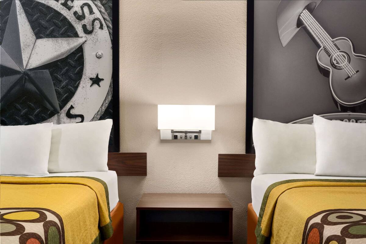 A hotel room with two beds, a small table, a wall-mounted light, and a guitar on the wall.