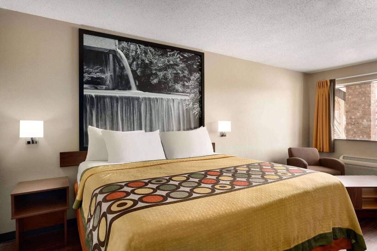 A hotel room with a large bed, two bedside tables, a chair, a window with curtains, and a wall-mounted picture.
