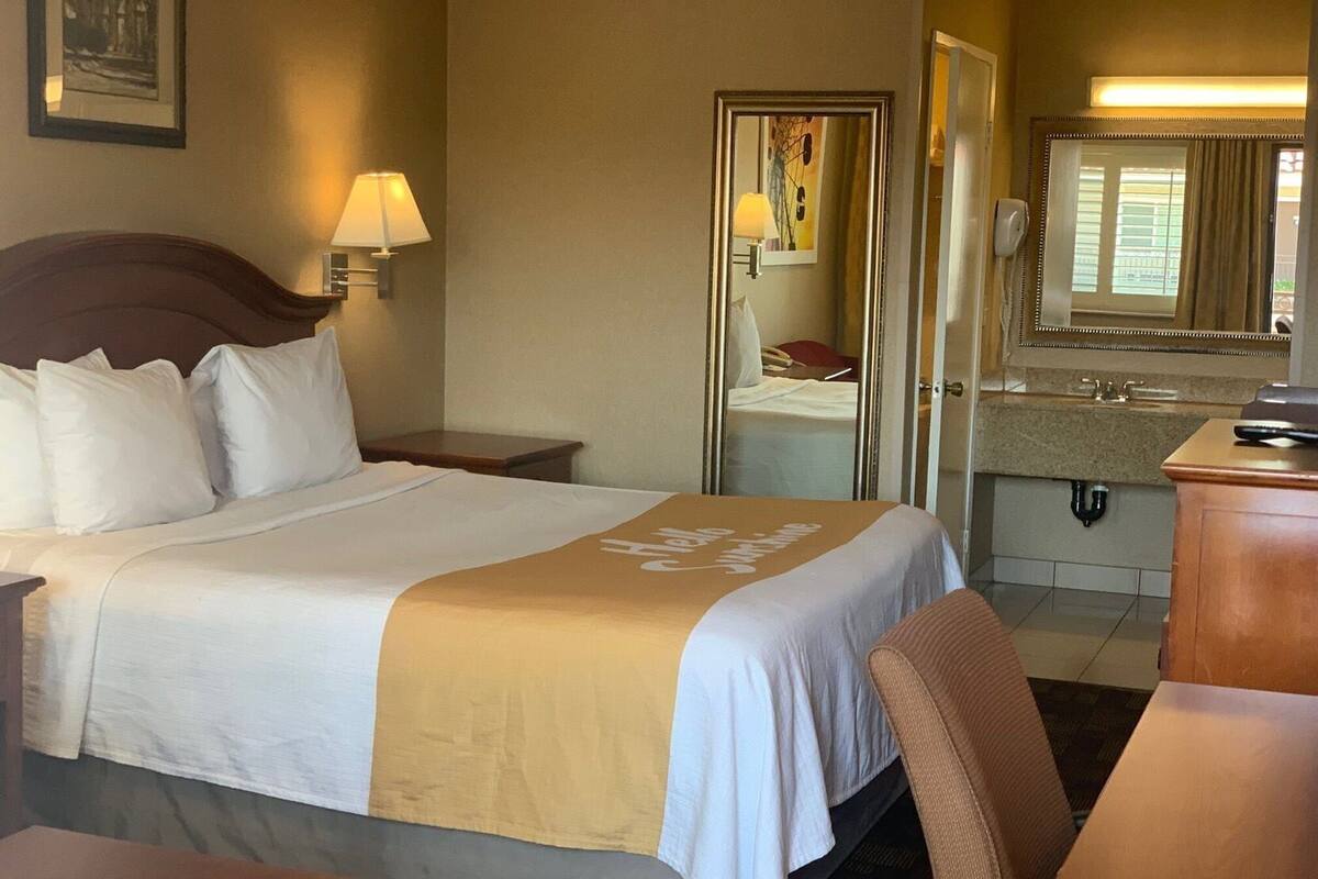 A hotel room with a bed, a desk, a chair, a mirror, a lamp, and a bathroom visible through an open door.