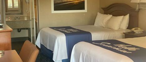 A hotel room with two beds, a bathroom, a desk, and a painting of a sunset.