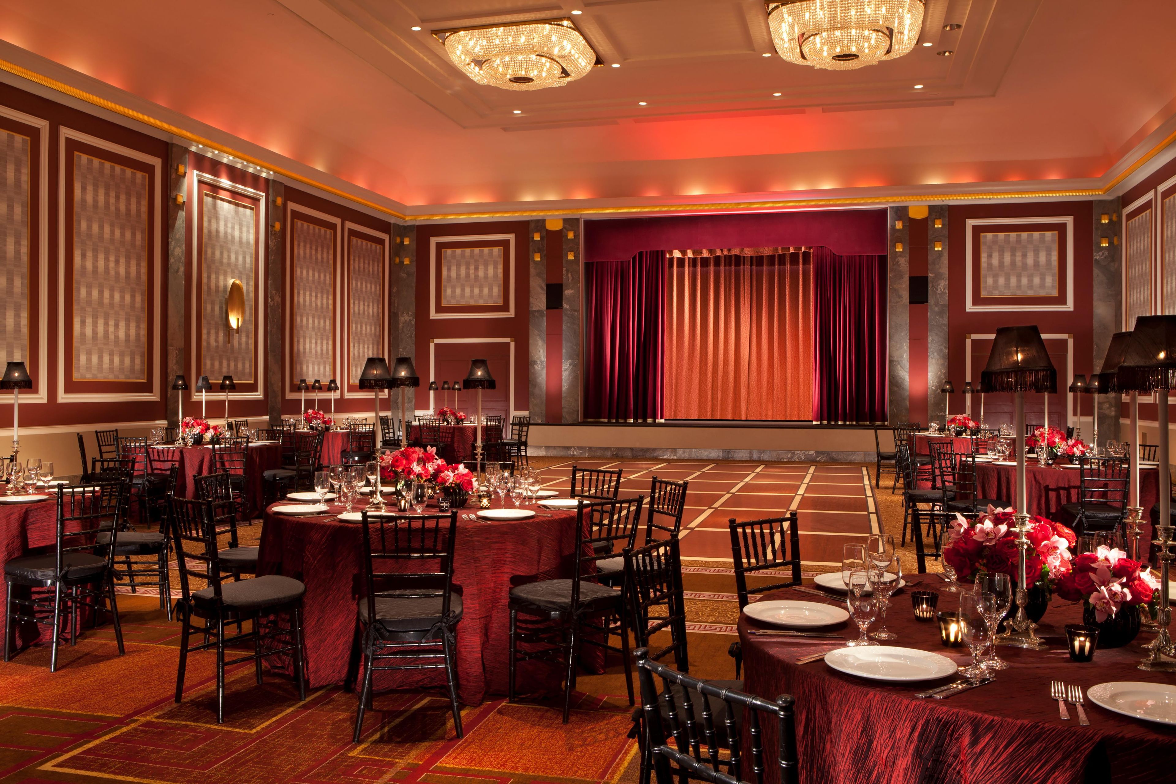 Un salón de banquetes con mesas redondas, sillas y un escenario con cortinas.