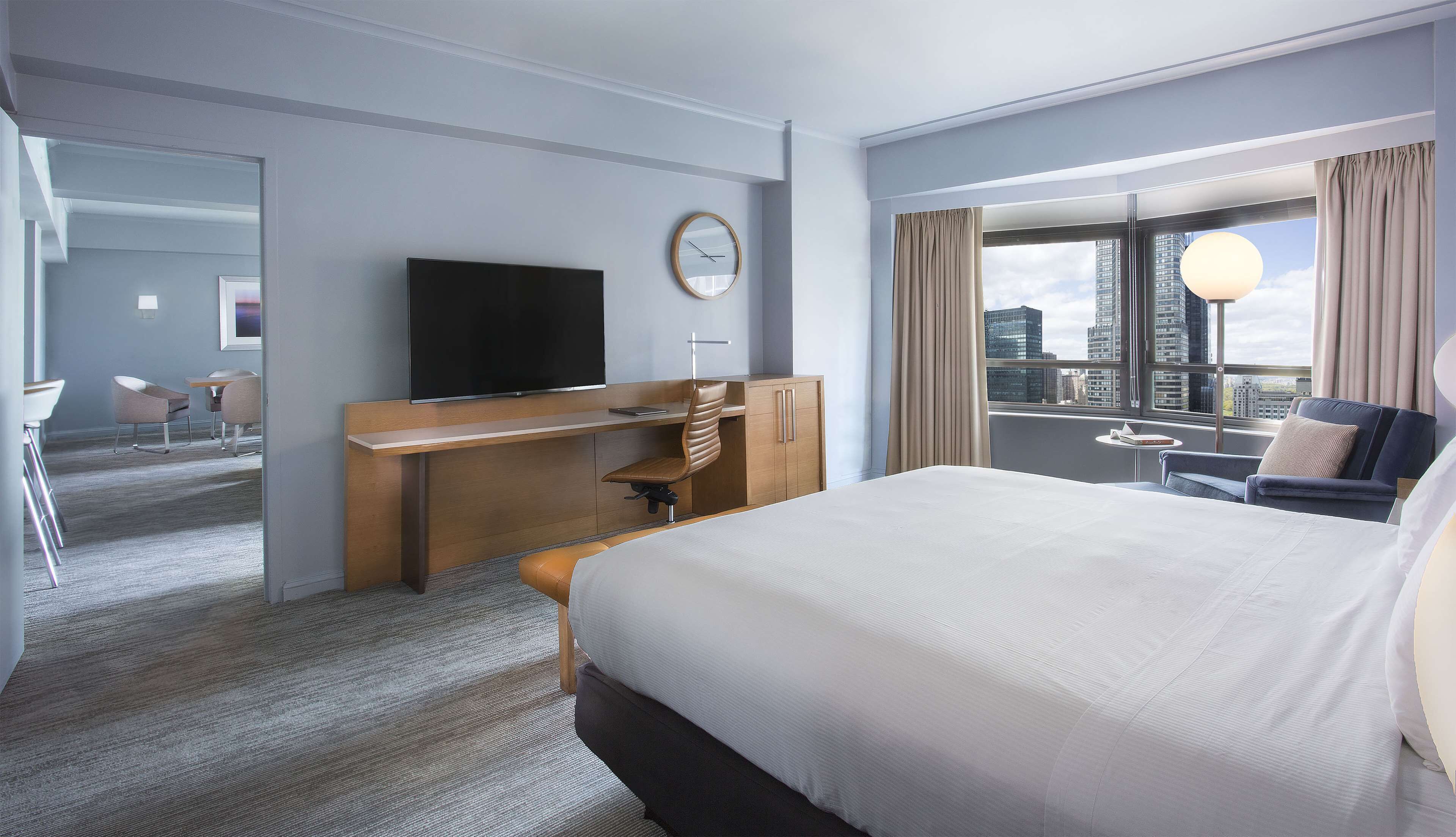 A hotel room with a large bed, a desk with a chair, a television, and a view of the cityscape through the window.