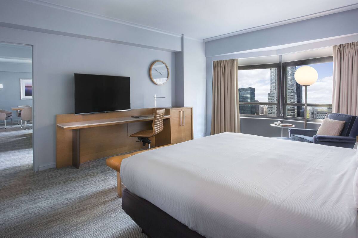 Hotellihuone, jossa on suuri sänky, työpöytä tuolilla, televisio ja ikkunasta avautuva näkymä kaupungin ylle.