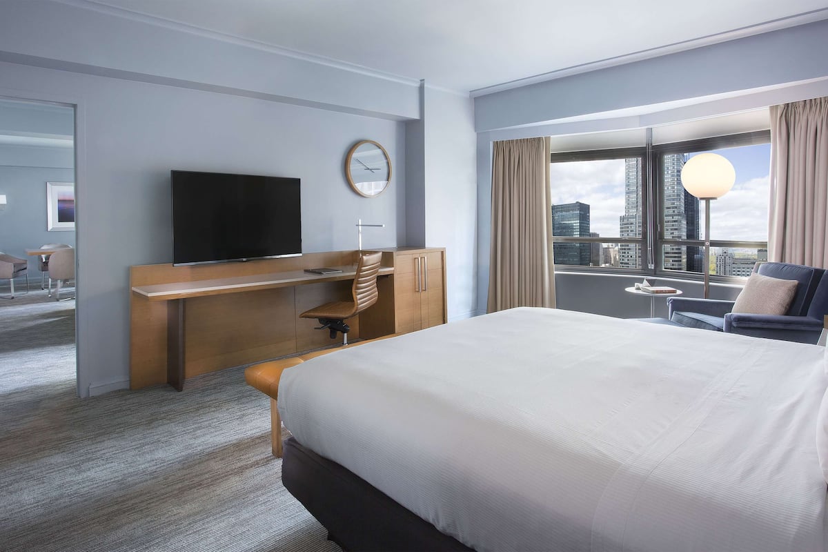 A hotel room with a large bed, a desk with a chair, a television, and a view of the cityscape through the window.