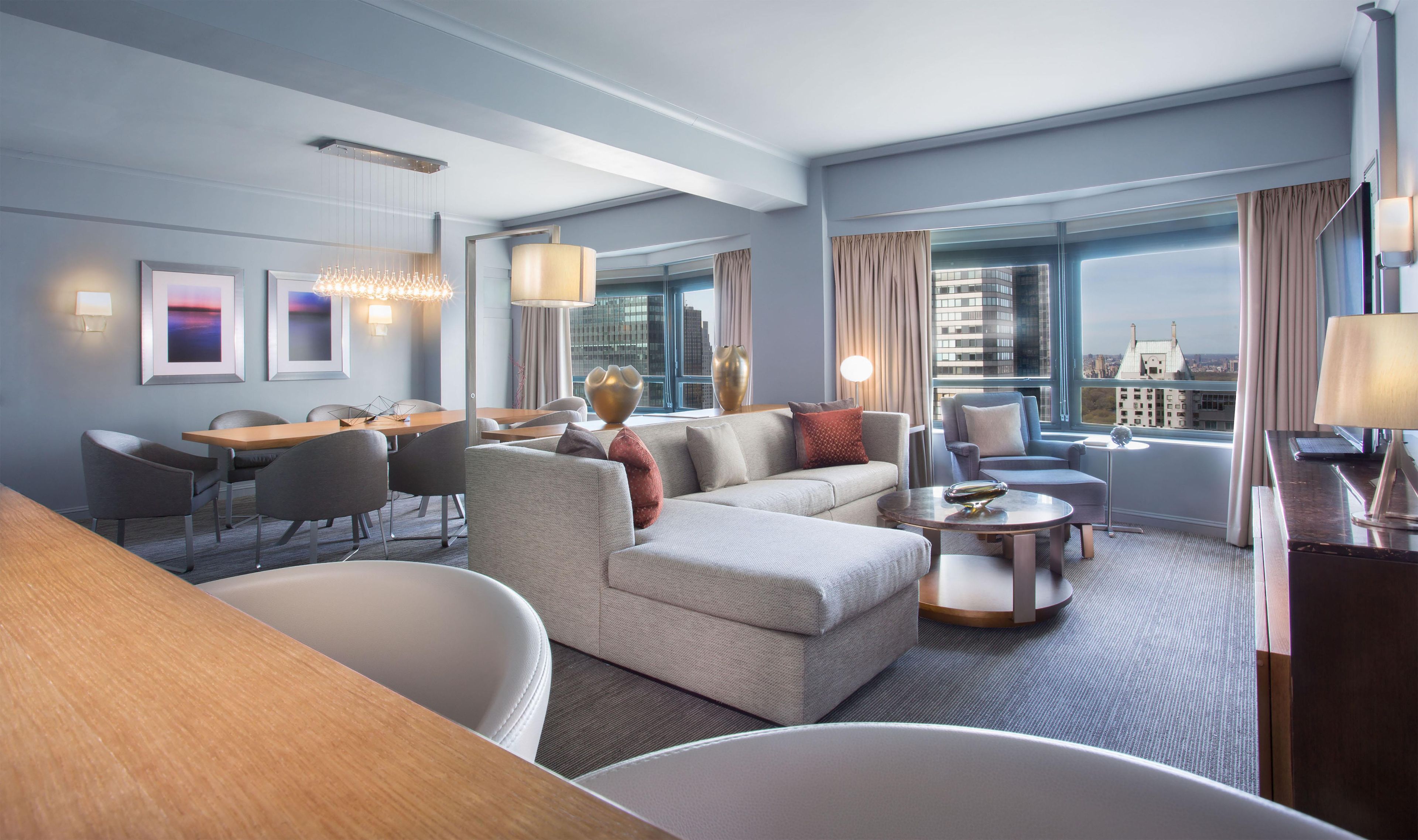 A modern hotel room with a dining area, seating, and a view of the cityscape.