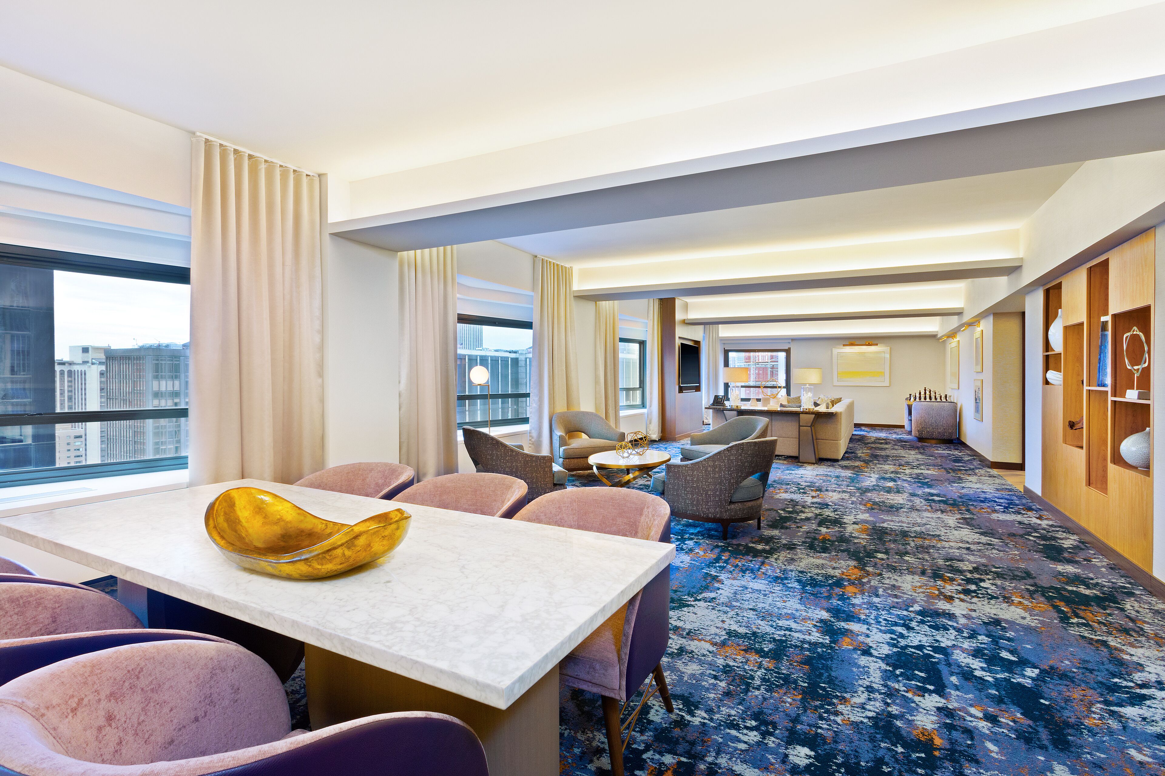 A hotel room with a dining area, a living area with a sofa and armchairs, and a large window offering a city view.