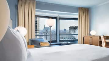 A hotel room with a large bed, a desk, a chair, and a view of a cityscape through a large window.