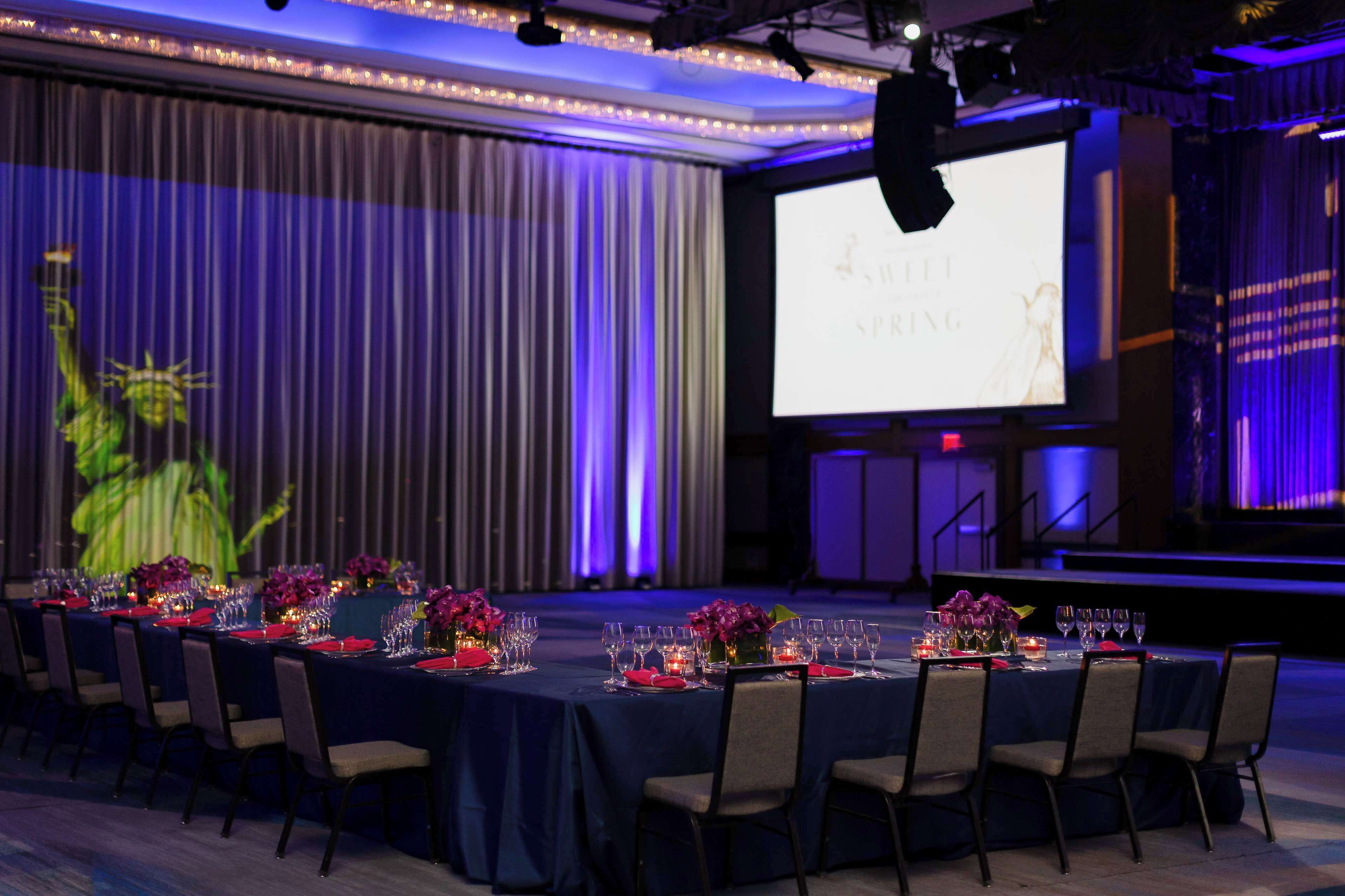 Un salón de banquetes con mesas dispuestas para un evento formal, una pantalla de proyección mostrando 'SILKET SPRING' y un escenario con un micrófono.