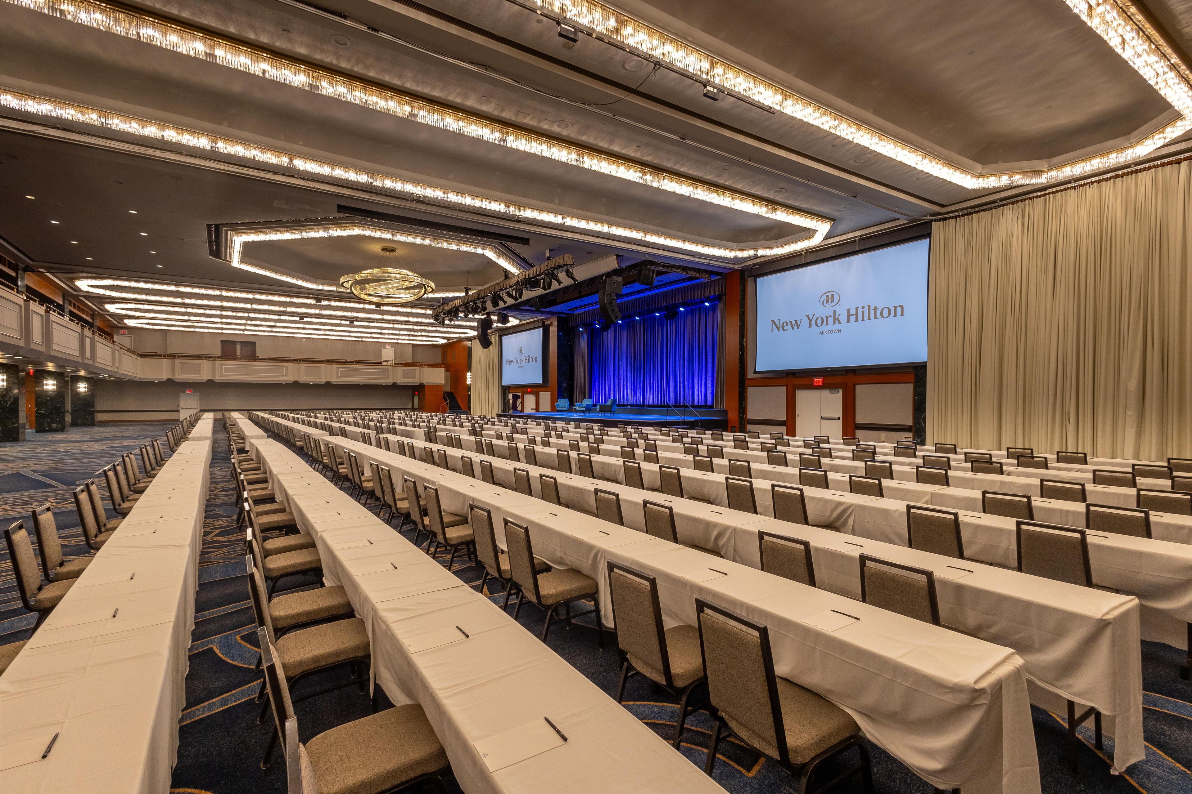 Un salón de conferencias con filas de mesas y sillas, un escenario con una pantalla de presentación y una araña de cristal.