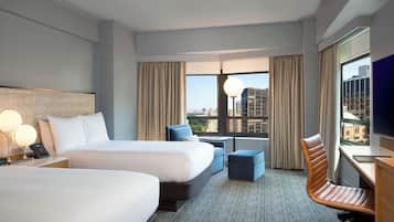 A hotel room with two beds, a desk, a chair, a lamp, and a view of the city.