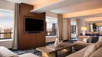 Una habitación de hotel moderna con un televisor de pantalla plana grande montado en un panel de madera, un conjunto de sofá color beige, una mesa de centro y vistas de la ciudad a través de amplias ventanas.
