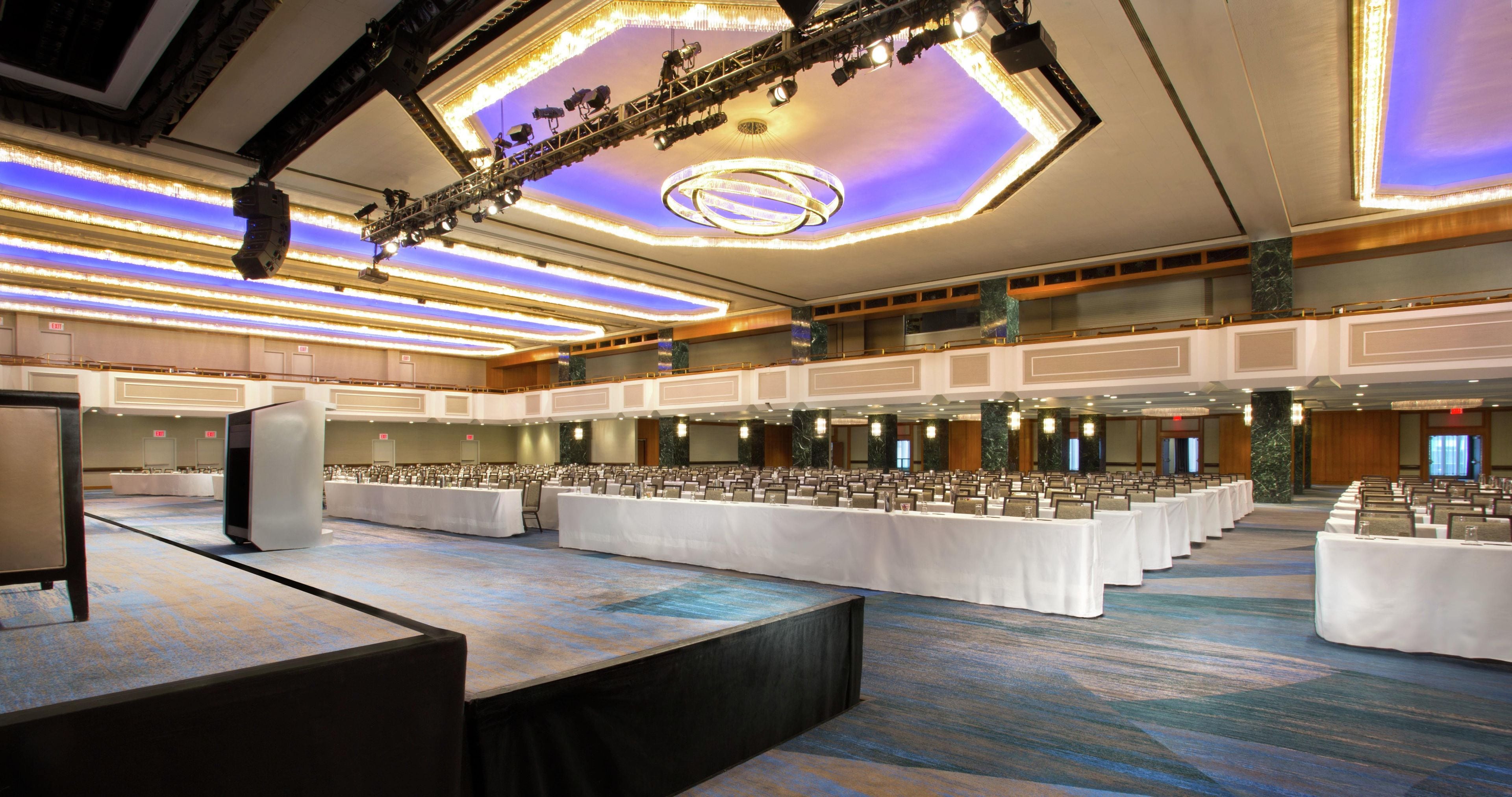 Une grande salle de conférence dotée de nombreuses tables et chaises, d’une scène équipée d’un système d’éclairage et d’un balcon donnant sur la salle.
