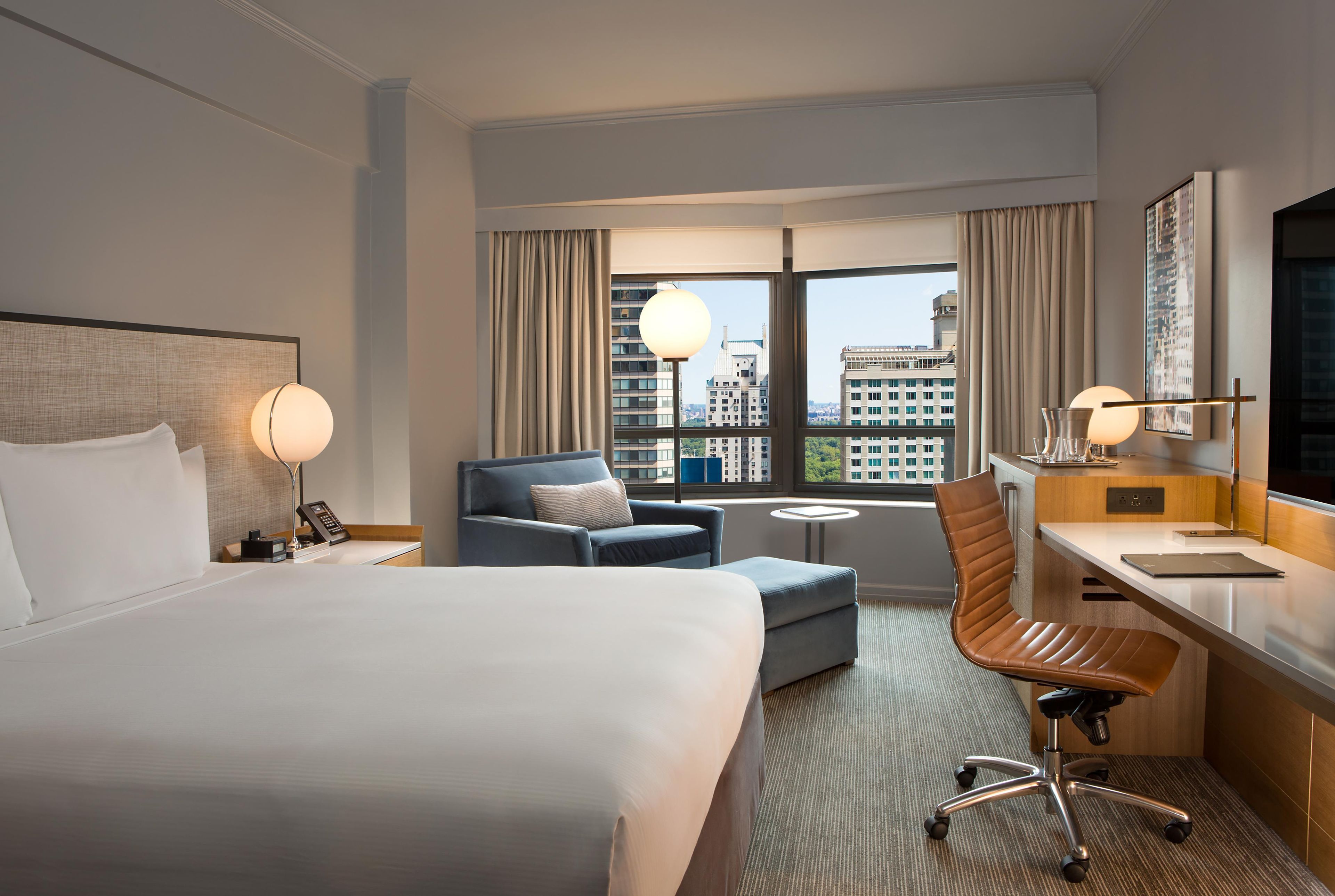 A hotel room with a large bed, a desk, a chair, and a view of the city through the window.