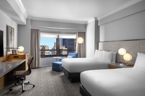 A hotel room with two beds, a desk, a chair, a TV, and a view of the city.
