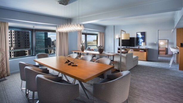 A modern hotel room with a dining area, a living area with a TV, and a view of the cityscape.