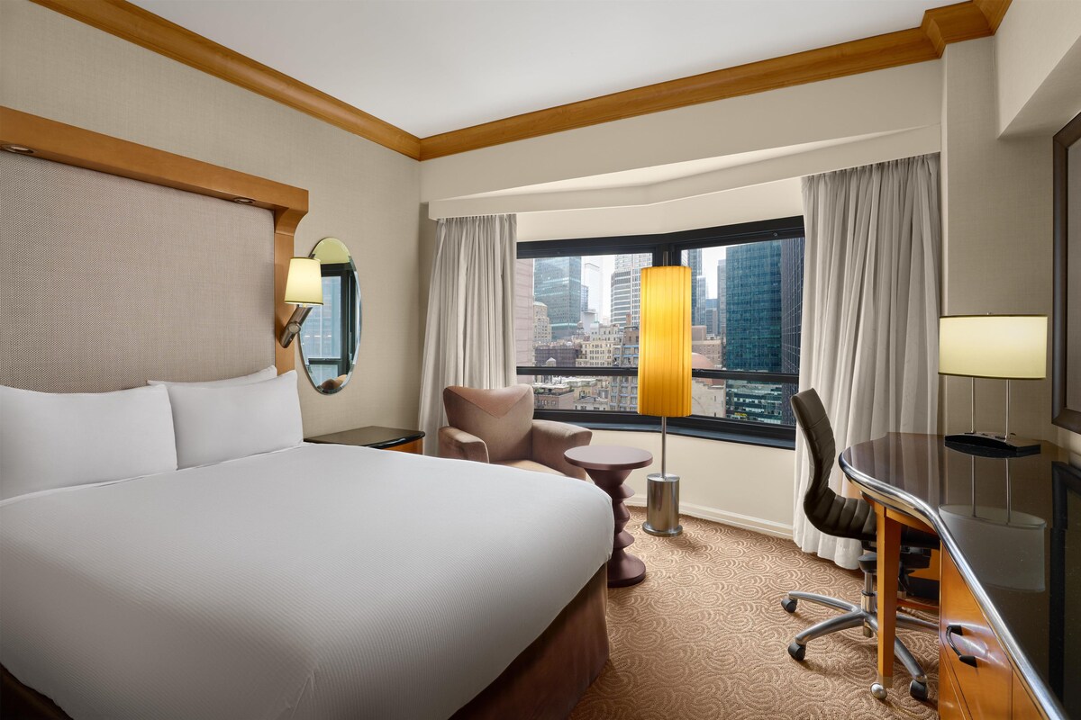 A hotel room with a large bed, a desk, a chair, a view of a cityscape, and a window with curtains.
