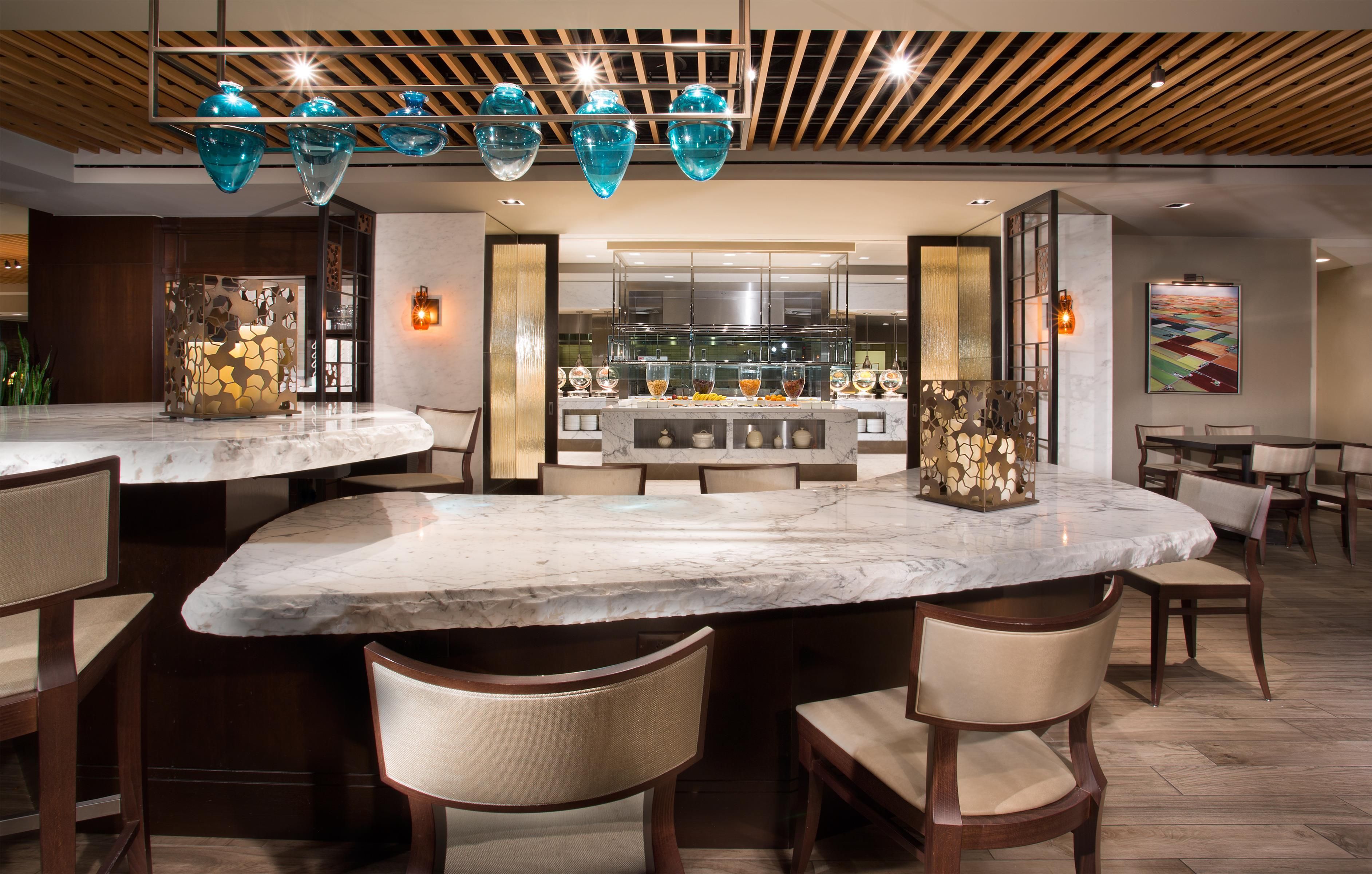 A modern bar area with marble countertops, hanging blue glass chandeliers, and a view of the kitchen in the background.