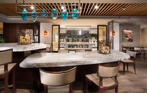 Zona de bar moderna con mostradores de mármol, lámparas colgantes de vidrio azul y vista a la cocina de fondo.