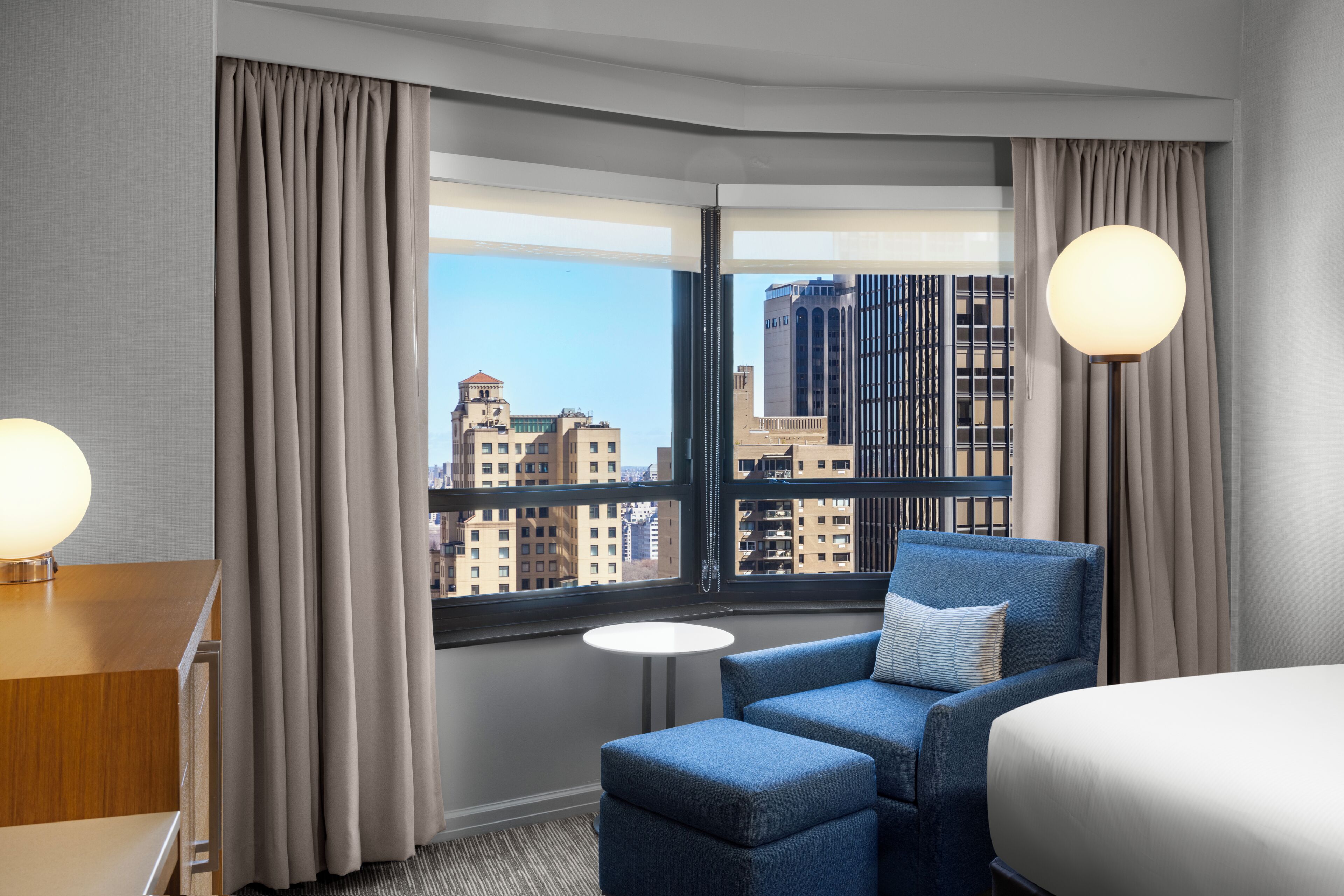 A hotel room with a large window, a blue armchair, a small round table, and a bed.