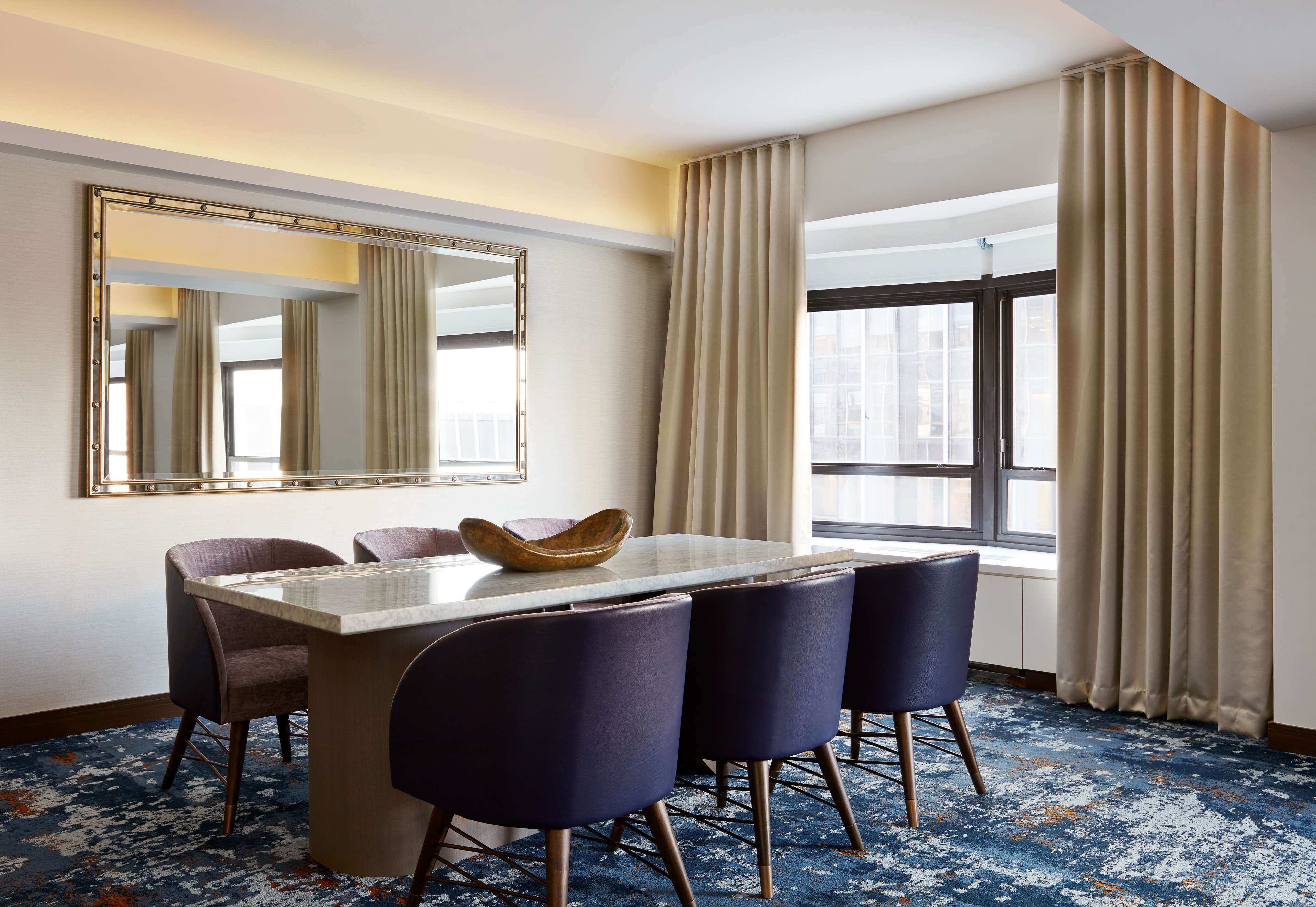 Une chambre d’hôtel dotée d’un coin repas comprenant une table en marbre, quatre chaises sombres, un grand miroir et une vue sur la ville, visible à travers une grande fenêtre ornée de rideaux beiges.