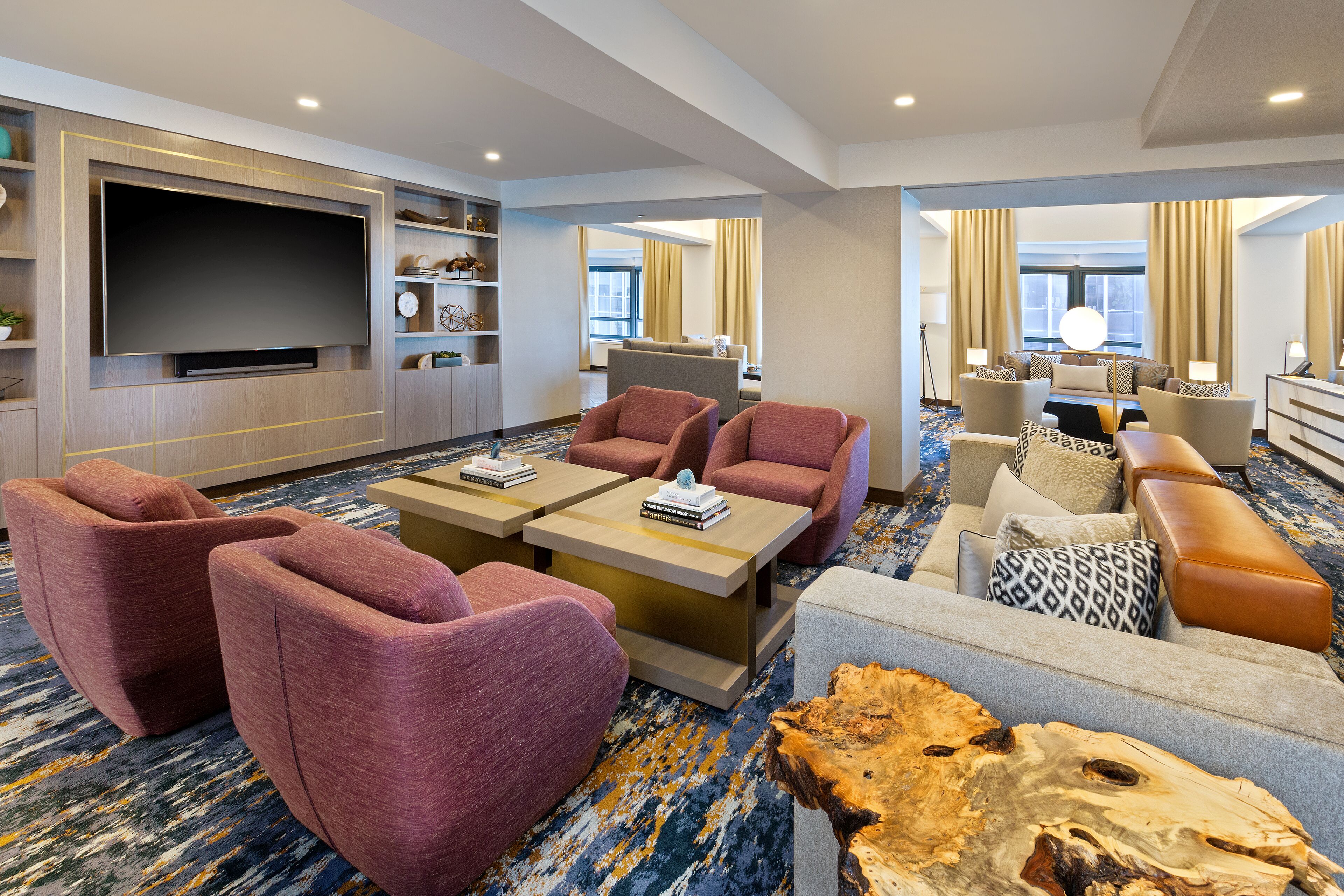 Habitación de hotel con un televisor de pantalla plana grande, una zona de estar con sofás y sillones, una mesa de centro y un ventanal con cortinas.