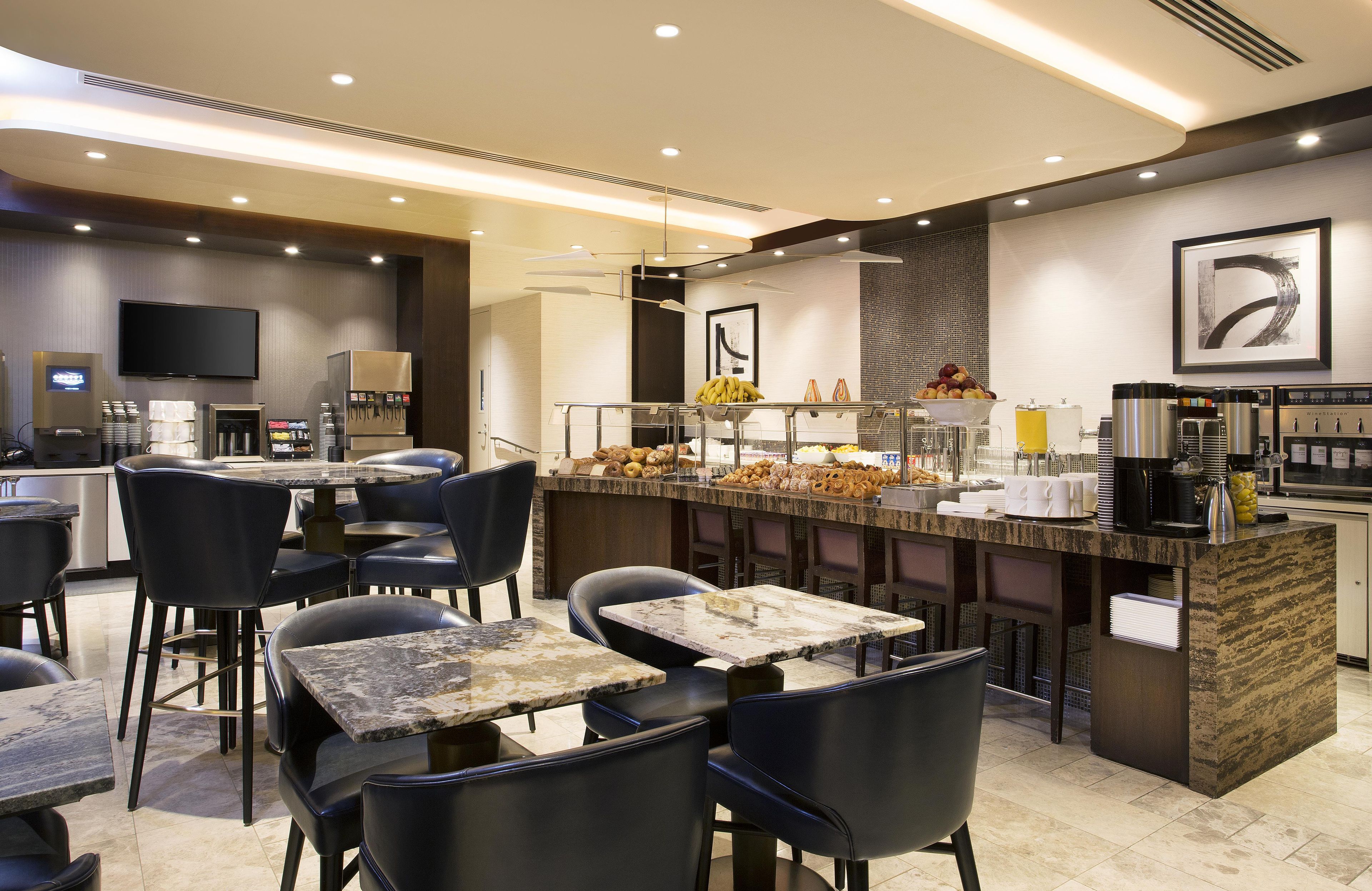 Un café moderne avec des tables en marbre et des chaises noires, un comptoir où sont exposés divers produits alimentaires, et un téléviseur à écran plat fixé au mur.