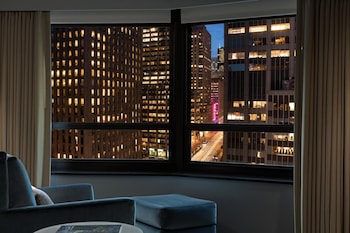 A hotel room with two beds, a desk, a chair, a TV, and a view of the city. at New York Hilton Midtown