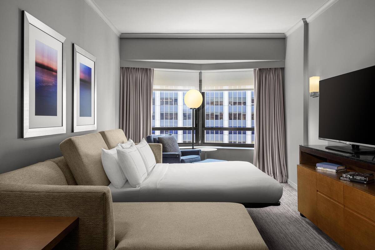 Moderni hotellihuone, jossa on suuri sänky, sohva, televisio ja suuret ikkunat, joista avautuu näkymä kaupunkiin.
