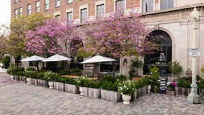 A brick building with a row of outdoor tables and chairs, surrounded by flowering trees and potted plants. - The Culver Hotel (Culver City)