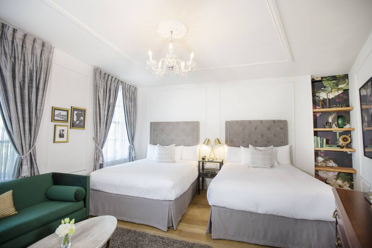 A hotel room with two beds, a green sofa, a bedside table with lamps, a chandelier, and a bookshelf.